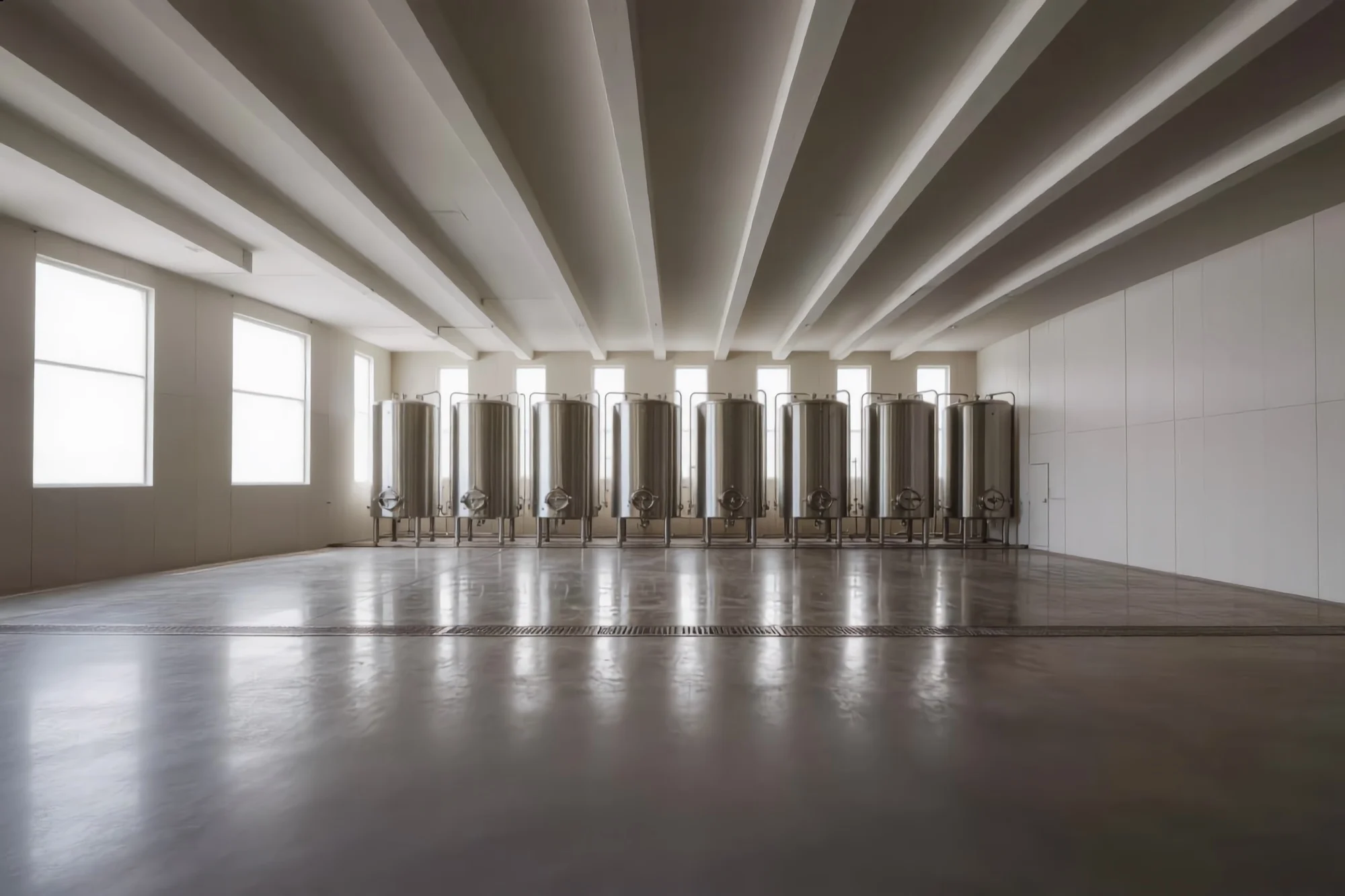 A seamless resin floor running unbroken beneath stainless tanks in a beverage production hall, a channel drain set flush in the surface